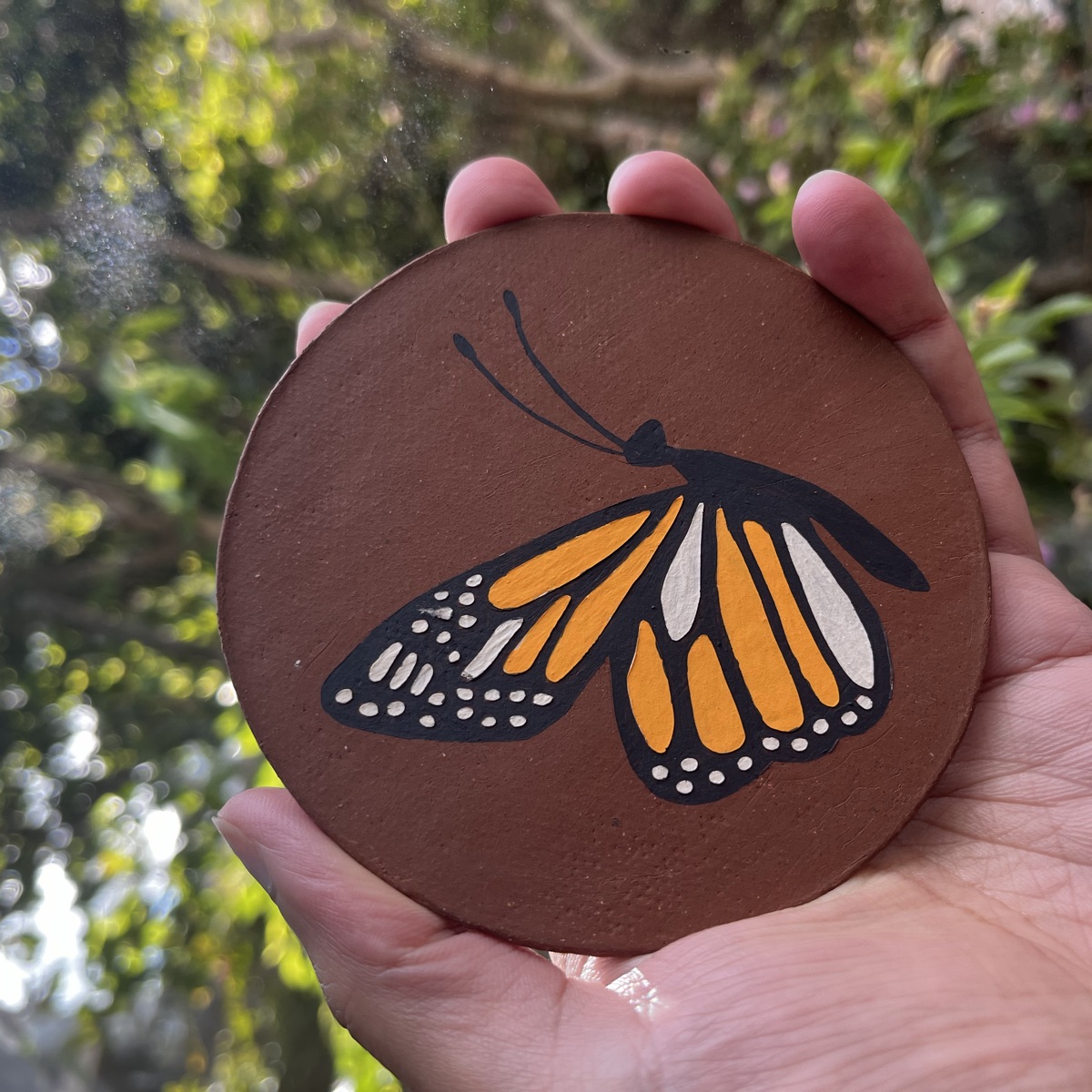 Round stoneware tile hand-painted with a monarch butterfly in orange, white and black, resting in an open palm