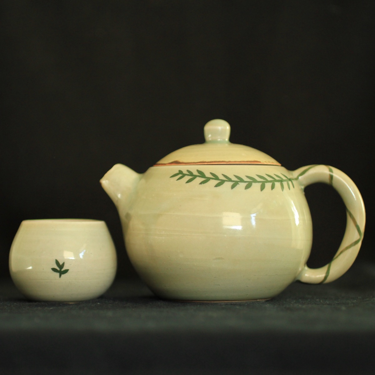 A small celadon-glazed teapot painted with a trailing green leaf vine, beside a matching cup with a single sprig, against a dark background