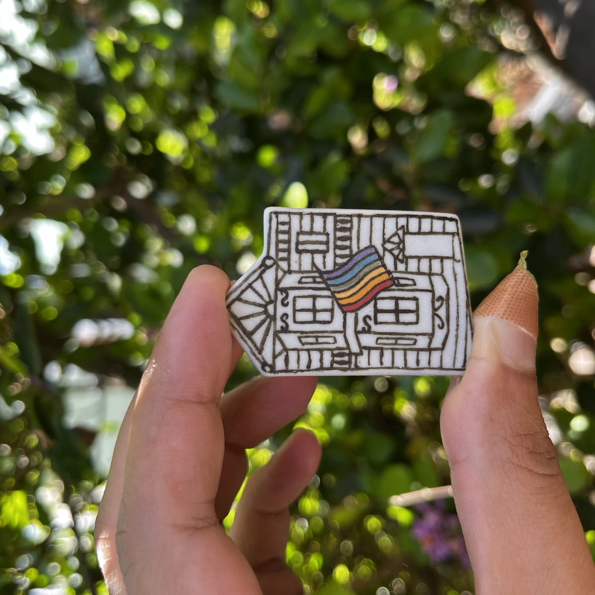 Small white ceramic tile shaped like a San Francisco Victorian house, with bay windows drawn in brown line work and a hand-painted rainbow flag above the door