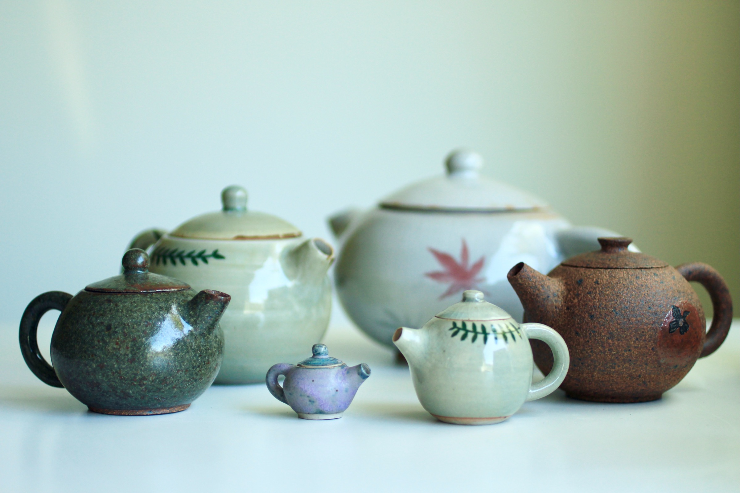 Six hand-thrown teapots arranged in a row, from a large white pot painted with a red maple leaf to a lilac miniature the size of a thumb